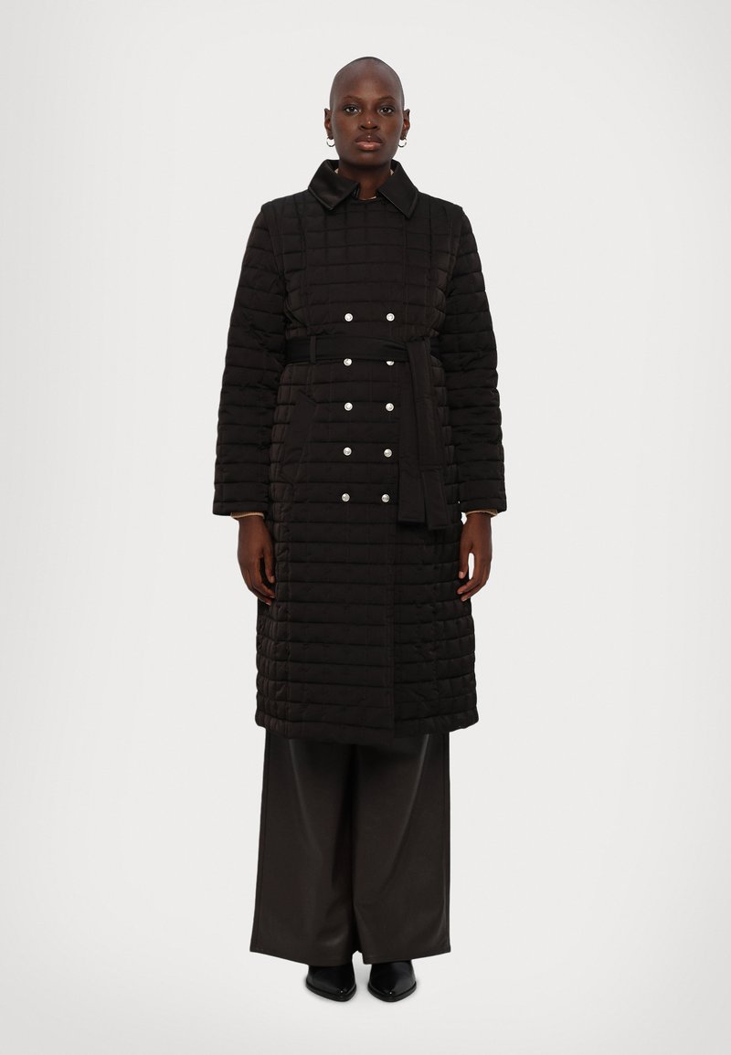 Melnā quiltota garā jaka ar jostiņu viduklī, sudraba pogu detaļām un apkakles izgriezumu. Iezīmē gludu tekstūru un taisnu piegriezumu.