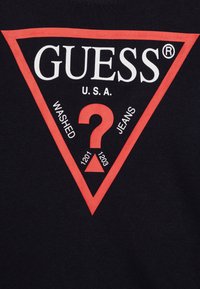 Zwarte t-shirt met een groot rood driehoekig logo met "GUESS U.S.A." en de tekst "WASHED" en "JEANS" in het wit, gecentreerd rondom een rode vraagteken.