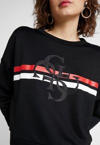 Sweat-shirt noir à col rond, avec un logo scintillant et des rayures horizontales rouges, blanches et noires sur la poitrine.