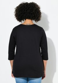 Schwarzes Shirt mit drei Viertelärmeln aus weichem Stoff, mit rundem Halsausschnitt und seitlichen Schlitzen, kombiniert mit blauen Jeans.