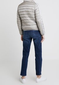 Gepolsterte, hellgraue Jacke mit horizontalem Steppmuster, hohem Kragen und reflektierenden Seitestreifen. Getragen mit dunkelblauen Jeans und weißen Sneakers.