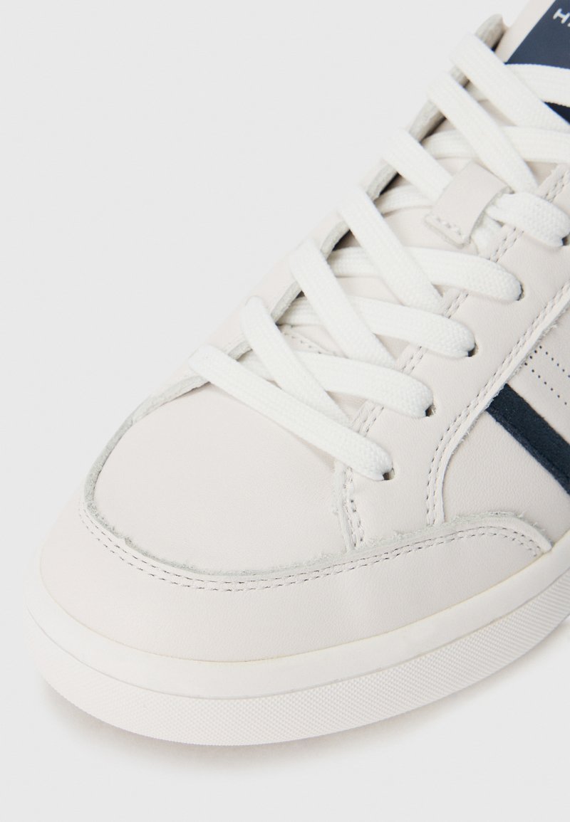 Gros plan sur l'embout d'une sneaker blanche avec des lacets blancs, des bandes noires sur les côtés et des coutures détaillées sur un fond blanc.