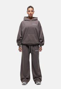 Overmaat grijze hoodie met een voorzak en wit logo, gecombineerd met ruime grijze joggingbroek. Zacht materiaal en een relaxte pasvorm.