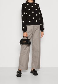 Schwarzer Pullover mit weißen Herzmustern, graue Hosen mit Nadelstreifen in weiter Beinform, schwarze Handtasche mit goldenen Beschlägen und klobige schwarze Slipper.