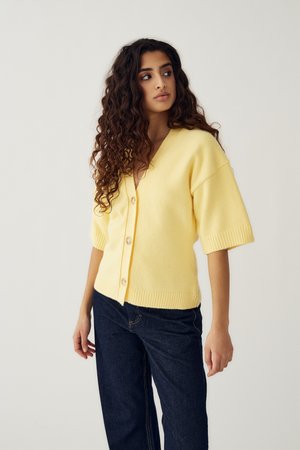 Mladá žena s dlhými kučeravými vlasmi, oblečená v svetložlte zapínanej kardigáne a tmavomodrých džínsach, pozerajúca sa do strany na jednoduchom pozadí.