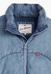 Denimjakke med høj krave, lys blå farve, lynlås foran og rød Levi's logoetiket på venstre bryst. Blød tekstur.