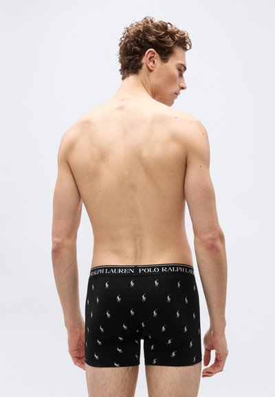 Άνδρας όρθιος με γυμνό κορμό, γυρισμένος πλάτη, φοράει μαύρα εσώρουχα boxer Polo Ralph Lauren με λευκό μοτίβο λογότυπου.