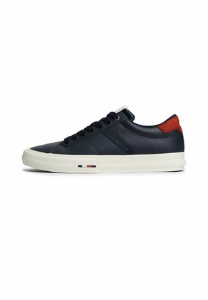 Niedriger schwarzer Sneaker mit weißer Sohle, schwarzen Schnürsenkeln, rotem Fersenlasche und kleinen rot-weiß-schwarzen Streifen an der seitlichen Sohle.
