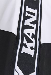 Juoda ir balta striukės detalė su dideliais užrašais ir kontrastingų audinių skyriais. Audinys atrodo lygus su siuvinėtais akcentais.