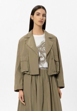 Frau mit langen, glatten Haaren trägt eine olivfarbene, kurze Jacke, ein weißes T-Shirt mit Aufdruck und einen passenden Rock mit hoher Taille und steht vor weißem Hintergrund.