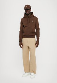 Sweatshirt castanho com capuz e bolso frontal, calças de linho bege e ténis brancos. Inclui também um boné castanho a condizer.