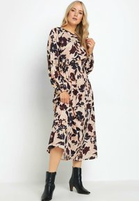 Blumenkleid mit langen Ärmeln in Hellrosa, mit dunkelvioletten und gelben Mustern, gerafftem Taillen-Design und schwingendem Rock. Getragen mit schwarzen Knöchelstiefeln.
