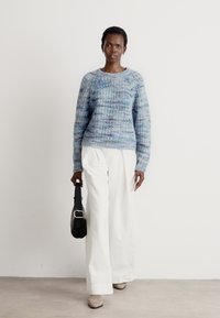 Blauer und lila gestrickter Pullover mit einem strukturierten Design, kombiniert mit weißen, weiten Hosen und einer schwarzen Schultertasche. Beigefarbene Stiefeletten.