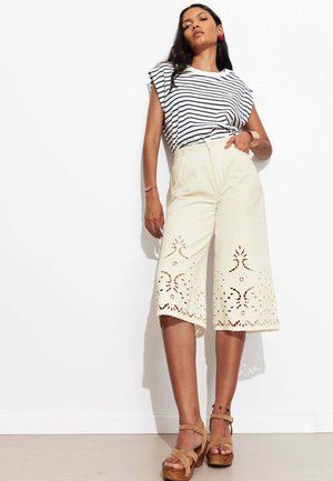 Femme portant un pantalon culotte blanc avec des motifs découpés au laser, un top sans manches rayé noir et blanc, et des sandales compensées beige, debout contre un mur blanc.