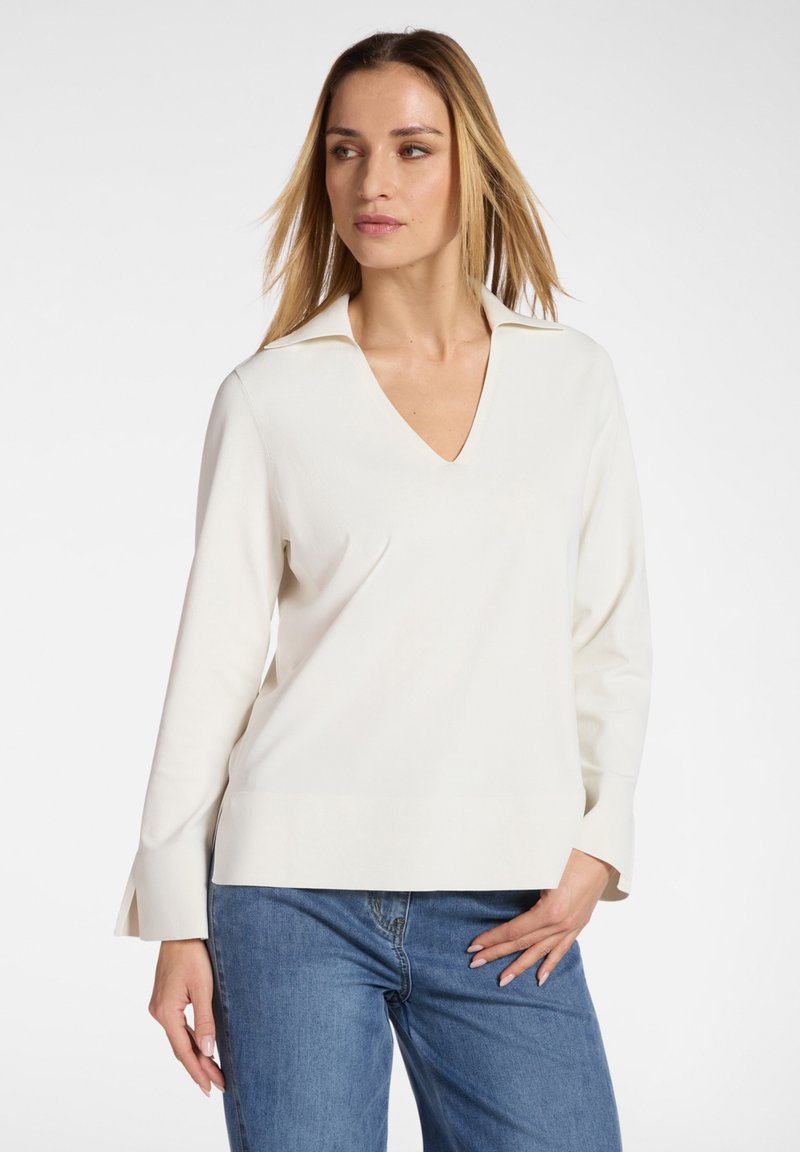 Donna che indossa una maglia bianca a maniche lunghe con scollo a V e collo, e jeans blu, posando con una mano sul fianco su sfondo bianco.