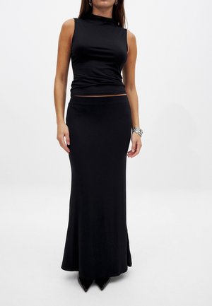 Woman wearing a sleeveless black top and long black skirt with black pointed shoes and a silver bracelet on right wrist.