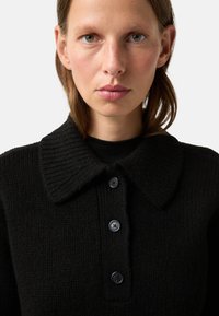 Black knitted sweater with a ribbed collar, featuring five buttons down the front. Close-up view highlighting texture and button details.