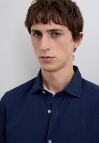 Camisa de botones azul marino con textura suave, que presenta un cuello puntiagudo y un sutil bordado de logo de calavera en el pecho.