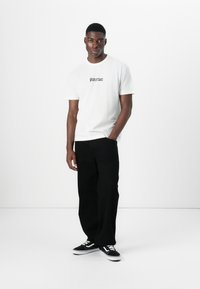 White cotton t-shirt with black graphic text, paired with black wide-leg trousers and black sneakers with white accents. Minimalist design.