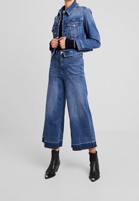 Jeansowa kurtka o klasycznym fasonie, jasnoniebieskim kolorze, z dwiema kieszeniami na piersiach i czarną bluzką pod spodem. Spodnie z szerokimi nogawkami z postrzępionymi brzegami oraz ciemne botki.