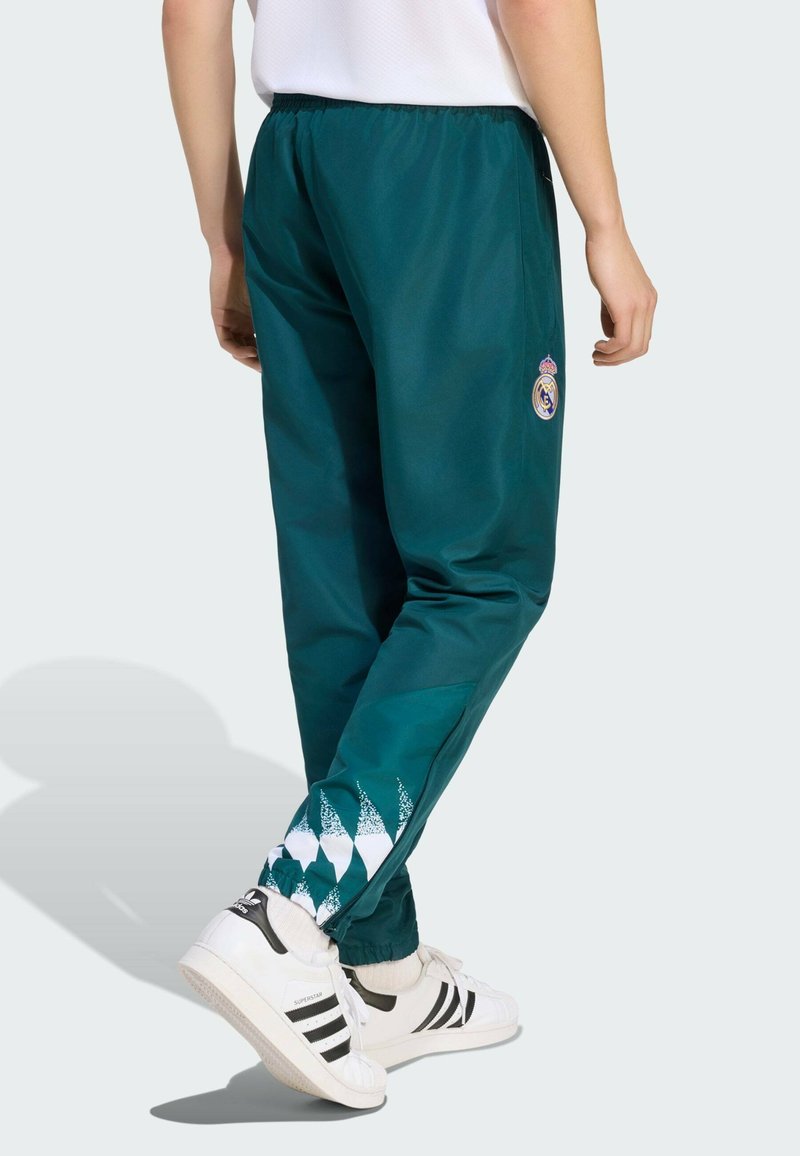 Person wearing dark green tracksuit pants with white and patterned lower legs and white Adidas sneakers, standing with one foot crossed behind the other.