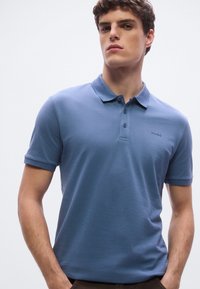 Polo krekls gaiši zilā kokvilnas maisījumā, ar apkaklīti, īsām piedurknēm un smalku logotipu uz krūtīm. Gluda tekstūra un piegriezums.