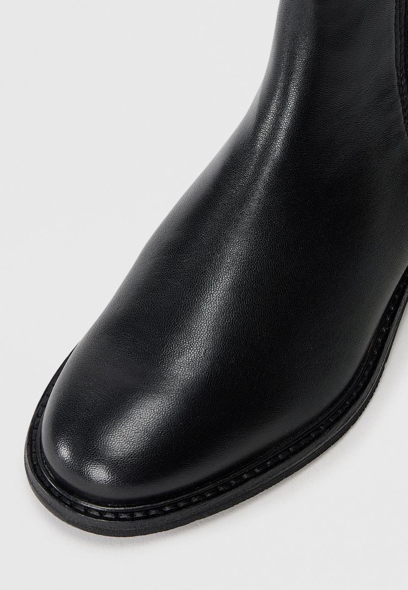 Schwarze Leder-Stiefelette mit glatter Textur, runder Spitze und kontrastierendem schwarzen Nähten entlang der Sohle. Minimalistisches Design ohne Verzierungen.