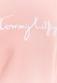 Tessuto di cotone rosa chiaro con logo "Tommy Hilfiger" ricamato in stile corsivo di colore bianco. Texture liscia con un sottile motivo a coste.