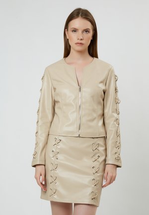 Veste en simili cuir beige avec fermeture éclair et détails lacés sur les manches, associée à une jupe assortie avec des accents en dentelle similaires.