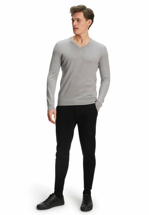 Maglione grigio a collo a V in materiale morbido a maglia, abbinato a pantaloni neri slim-fit e sneaker nere, in piedi contro uno sfondo bianco.