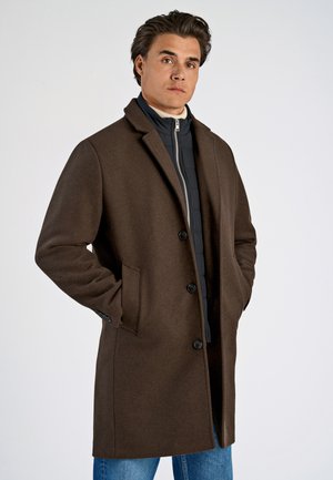 Cappotto di lana marrone con un design sartoriale, apertura con bottoni, tasche laterali e colletto classico. Indossato sopra un giubbotto imbottito nero.