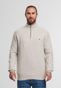Hellbeiger Pullover mit hohem Kragen und Reißverschluss, der ein strukturiertes Strickmuster und ein kleines dunkles Logo auf der linken Brust aufweist.
