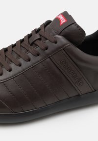 Sneaker in pelle marrone con lacci piatti marroni, pannelli laterali texturizzati e suola in gomma. Presenta un'etichetta rossa con logo sulla linguetta.
