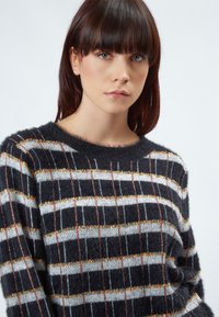 Gestickter Pullover mit flauschiger Textur, der schwarze, weiße und metallische Streifen aufweist. Rundhalsausschnitt mit mehrfarbigen horizontalen Mustern.
