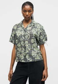 Fonott hajú nő rövid ujjú, gombos ingben, szürke és zöld virágos kockás mintával, és fekete nadrágban, egyszínű háttér előtt.