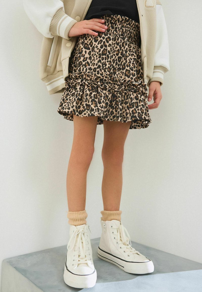 Child wearing a leopard print ruffled skirt, black top, beige jacket, beige socks, and white high-top sneakers standing on a gray platform.
