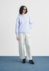 Γυναίκα που φοράει ανοιχτό μωβ φούτερ New Balance, γκρι φόρμα, μαύρα αθλητικά παπούτσια και μαύρα ορθογώνια γυαλιά ηλίου, στέκεται σε μπλε μοκέτα.