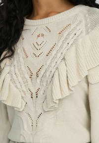 Sweter robiony na drutach w kremowym kolorze z teksturowanym wzorem, ażurowym designem i falbankami na ramionach dla dodatkowego akcentu.