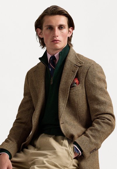 Blazer em espinha de peixe castanho sobre um suéter verde-escuro com fecho éclair. Camisa rosa-clara e gravata riscada visíveis. Lenço de bolso com padrão xadrez. Calças bejes.