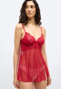 Vrouw draagt rode kanten en mesh doorzichtige lingerie jurk met dunne bandjes, kijkt iets naar de zijkant tegen een effen lichte achtergrond.