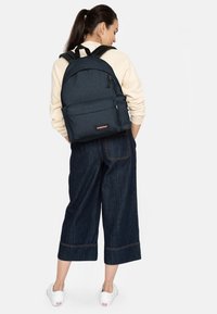 Mochila azul oscura con correas de hombro acolchadas, bolsillo frontal y una superficie texturizada. Llevada con un suéter claro y jeans oscuros de pierna ancha.