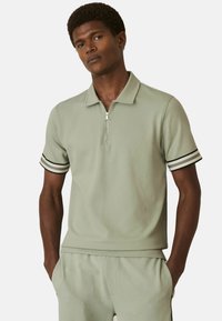 Tricou polo din țesătură texturată de culoare verde deschis, cu fermoar scurt, având accente în dungi negre și albe pe mâneci și guler.