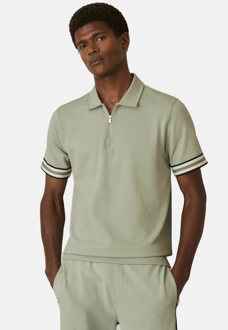 Tricou polo din țesătură texturată de culoare verde deschis, cu fermoar scurt, având accente în dungi negre și albe pe mâneci și guler.