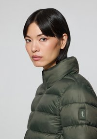 Puffig grön jacka med hög krage och dragkedjestängning, med en texturerad look och en logotyp på vänster ärm.