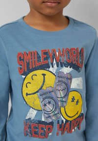 Blaues Langarm-T-Shirt mit gelben Smile-Gesicht-Grafiken, mehrfarbigen Ticketdesigns und dem Text "SMILEYWORLD KEEP HAPPY".