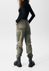 Pantalons cargo verts avec un patchwork, deux poches latérales et des poignets retroussés, associés à des bottes noires à lacets et un haut noir ajusté à manches longues.