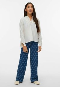 Camicia bianca a maniche lunghe con tessuto strutturato e scollo a V, abbinata a pantaloni di denim blu con motivo a cuore, completata da scarpe chiare.