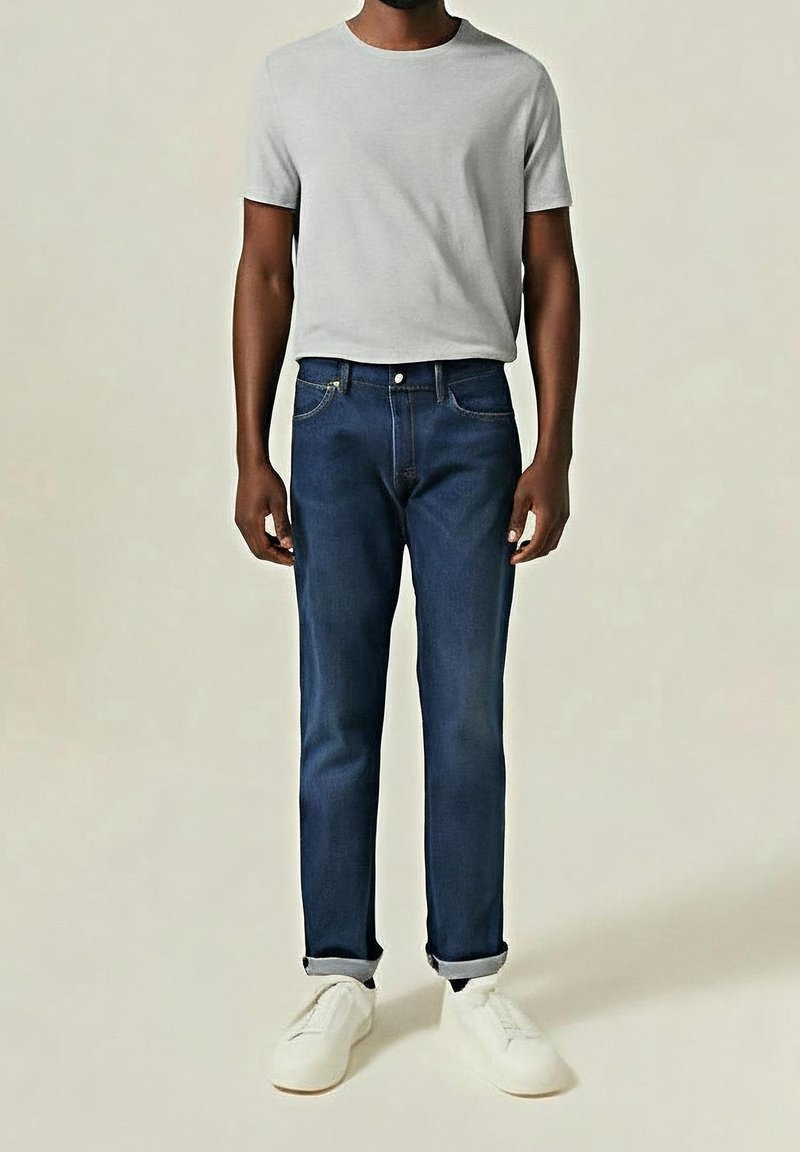 Homme portant une chemise à manches courtes gris clair, un jean bleu foncé avec des revers roulés et des baskets blanches, debout devant un fond clair uni.