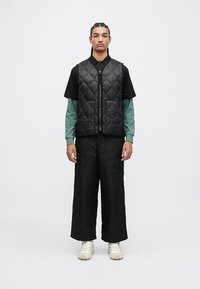 Jovem com cabelo trançado, vestindo um colete acolchoado preto e calças, sobre uma camisola polo preta e uma camisa de mangas longas verde, com ténis brancos.