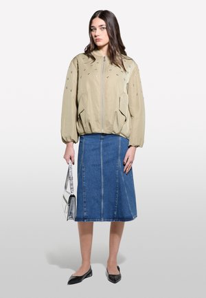 Veste bomber beige clair avec des embellissements métalliques, associée à une jupe en denim arrivant aux genoux et des ballerines noires. Sac à main avec des accents argentés.